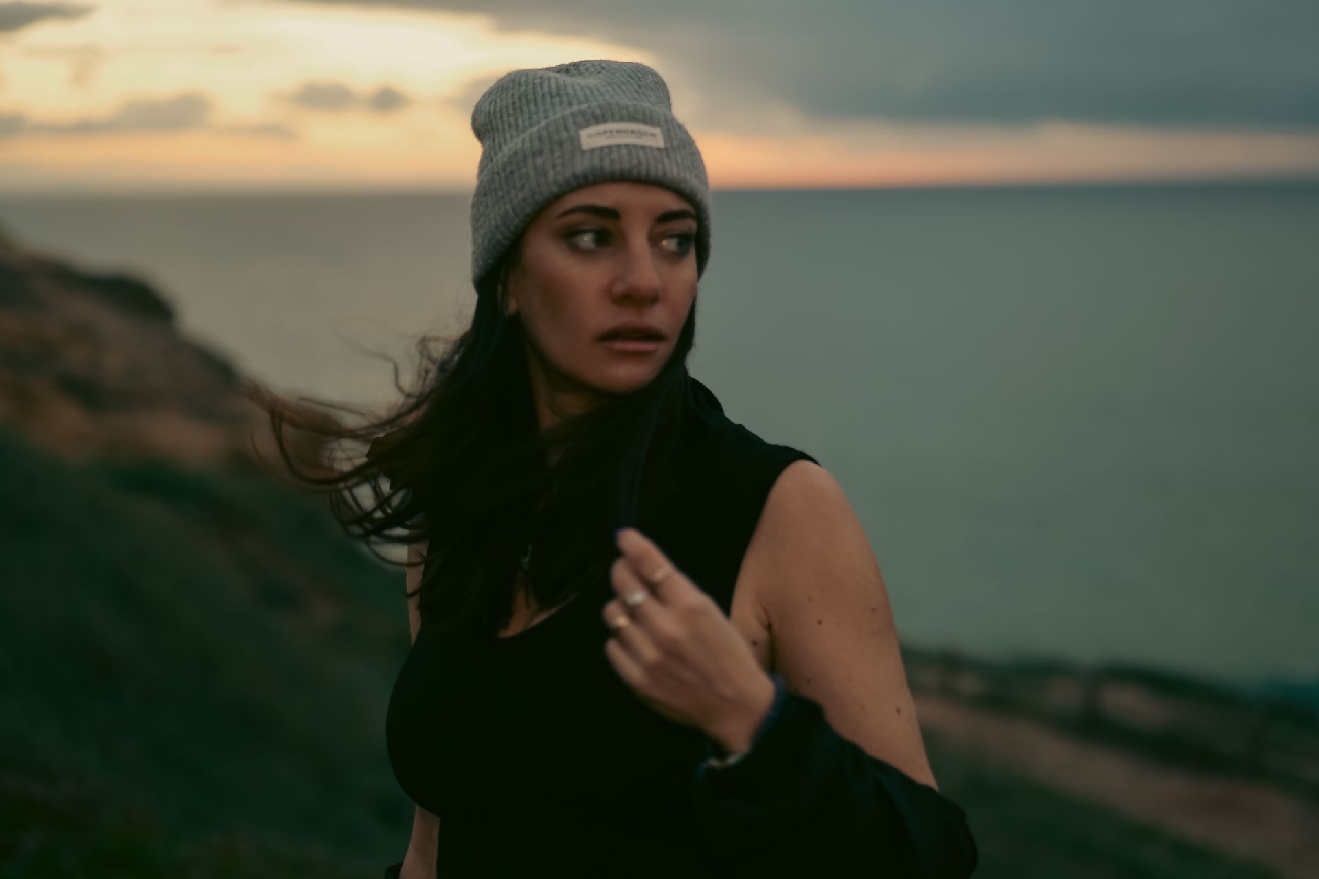 Woman in beanie standing by the sea at sunset, looking to the side.