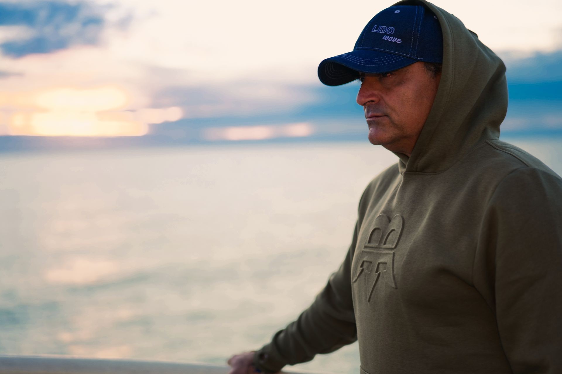 Person wearing a hoodie and cap, standing near a body of water during sunset.