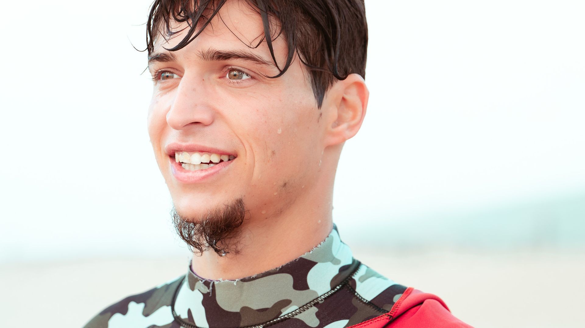 Smiling young man with wet hair wearing a camouflage-patterned wetsuit on a beach.