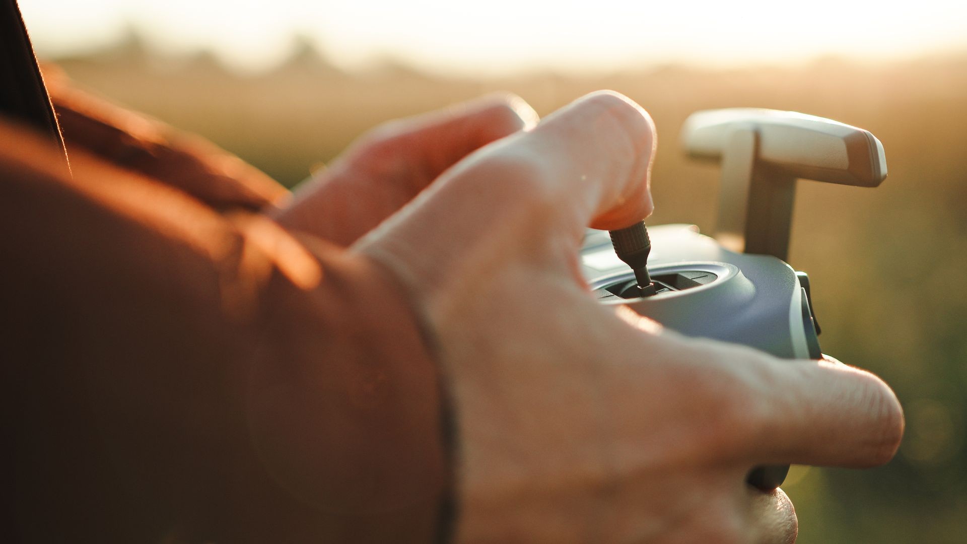 Close-up of hands operating a drone controller with a grassy field in the background during sunset.